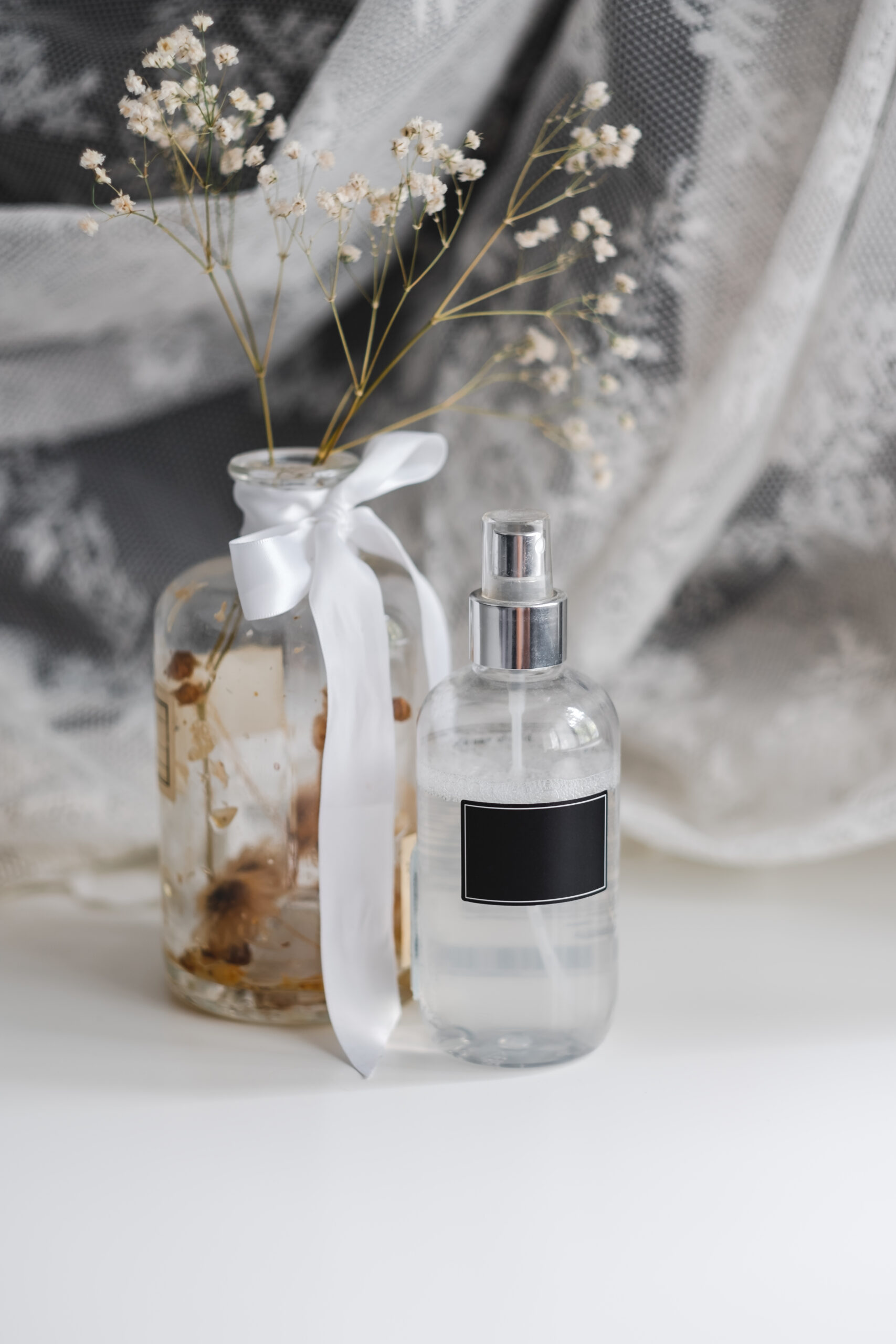 Fragrance spray in a transparent plastic bottle with black empty tag and dried white flowers in a vase, lace fabric.