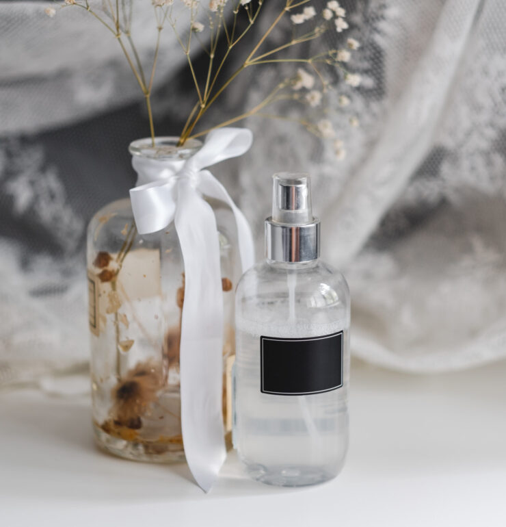 Fragrance spray in a transparent plastic bottle with black empty tag and dried white flowers in a vase, lace fabric.