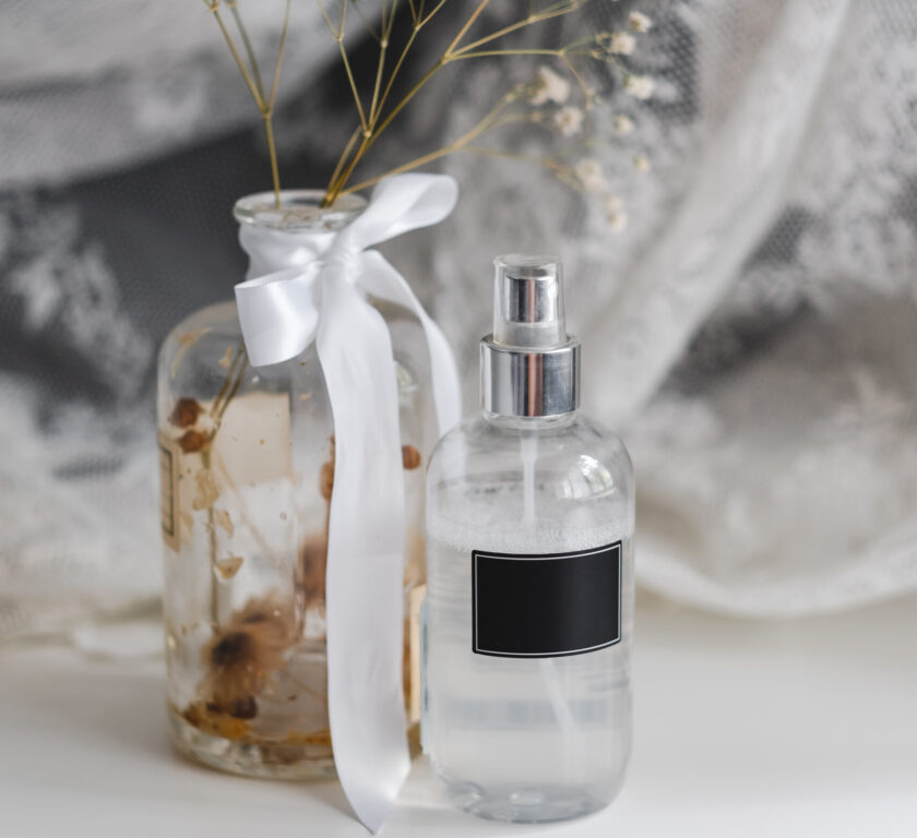 Fragrance spray in a transparent plastic bottle with black empty tag and dried white flowers in a vase, lace fabric.